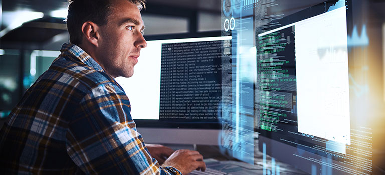 Man sitting in front of two desktop screens, parsing through code and data